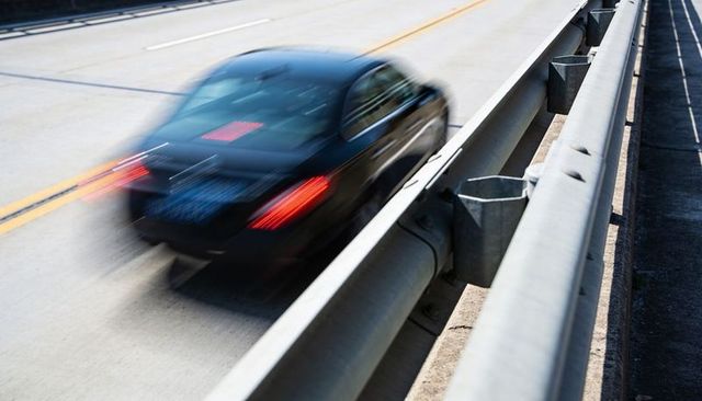 Speeding dark sedan creating motion blur and red taillight streaks on sunlit highway guardrail