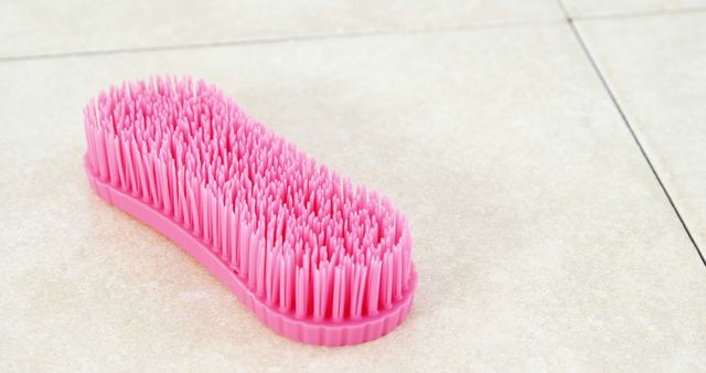 A pink scrub brush lies on a tiled surface, with copy space. Its bristles and ergonomic design suggest it's used for cleaning tasks around the home or workplace.