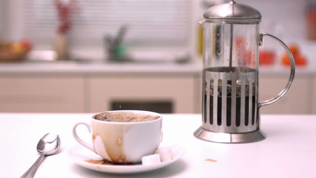 Close-up view of a sugar cube creating a splash as it drops into a coffee cup placed on a clean kitchen counter. Beside the cup is a stainless steel French press containing freshly brewed coffee. This dynamic scene captures the excitement and allure of morning routines and can be used in advertising coffee products, kitchenware, or demonstrating culinary aesthetics. Ideal for use in lifestyle blogs, cafe promotions, or recipe digital publications.