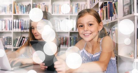 Smiling schoolgirl studying with tablet and laptop in modern library with bokeh lights