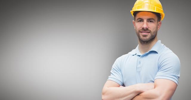 Construction worker wearing a yellow hard hat and light blue polo shirt standing with arms crossed against a grey background. Ideal for use in industrial, engineering, manual labor, and construction-related advertising or educational materials.