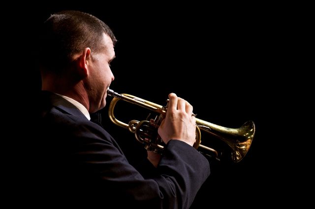 Musician in a suit performing skillfully on a trumpet against a dark background. Suitable for content related to musical performances, live concerts, professional musicians, and artistic expression in music.