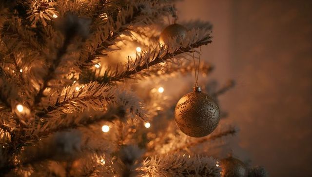 Gold christmas bauble hanging on flocked tree branch with warm bokeh lights, cozy holiday