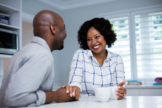 This image depicts a happy couple enjoying a moment together in their kitchen while having coffee. They are smiling and holding hands, showcasing a strong bond and affection. This image can be used in advertisements, blogs, or articles related to relationships, home life, morning routines, or coffee brands.
