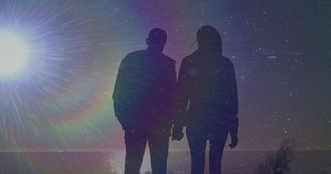 Silhouette couple holding hands facing ocean horizon at night with colorful lens flare