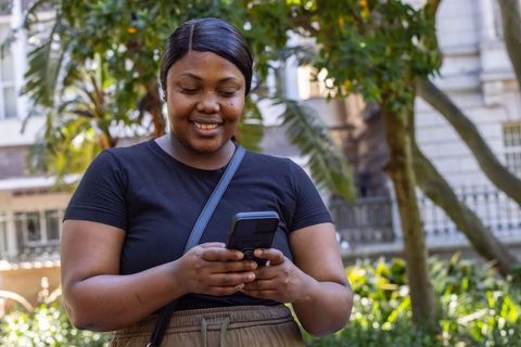 African american woman texting on smartphone, smiling in sunlit urban courtyard