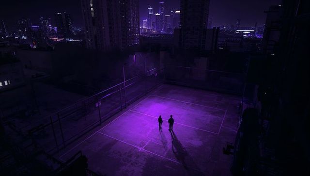 Two silhouettes standing on neon-lit rooftop court watching city skyline at night