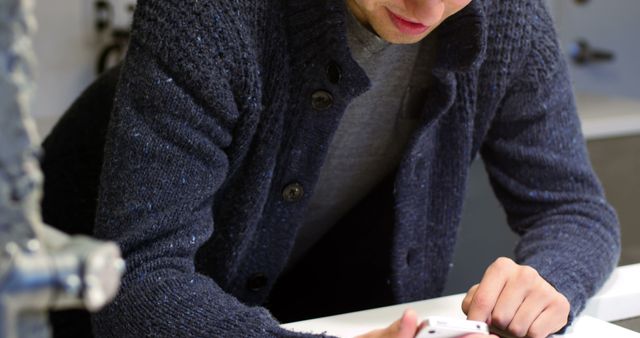 A man dressed in a casual blue knitted sweater is using a smartphone while leaning over a white surface in an indoor urban environment. Perfect for themes related to modern technology, casual fashion, daily lifestyle, or connectivity.