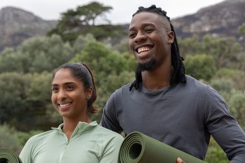 Smiling diverse couple holding yoga mats on trail practicing outdoor wellness and yoga