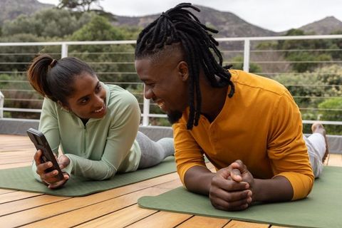 Diverse couple practicing yoga and sharing smartphone on outdoor terrace deck at sunrise