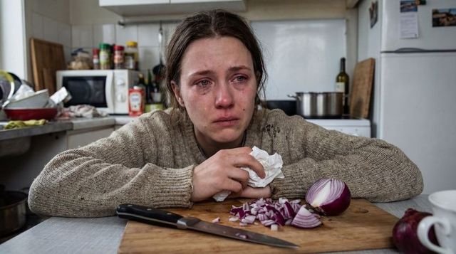 Young woman crying in cluttered kitchen holding tissue beside chopped onion and knife