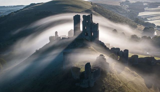 Misty dawn flowing fog around medieval castle ruins on hilltop, backlit countryside scene