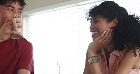 Smiling interracial couple sharing coffee and conversation near blinds with smartwatch