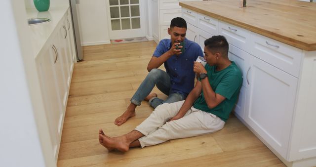 Two men sit on a kitchen floor together while sharing a warm drink, signifying a moment of relaxation and friendship. Useful for illustrating themes of companionship, casual home life, or weekend leisure activities.