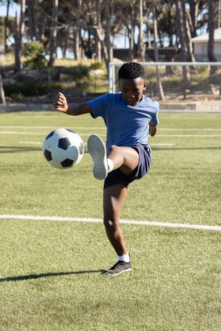 Teenage african american boy kicking soccer ball on sunlit turf near goal, athletic action