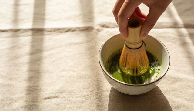 Hand whisking vibrant matcha in ceramic bowl on sunlit beige linen with frothy foam