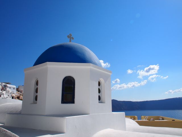 Rounded white building with a blue dome and a cross on top overlooking a vast sea under a bright, sunny sky. Iconic Greek architecture making it perfect for travel brochures, cultural tours advertisements, or summer vacation promotions.