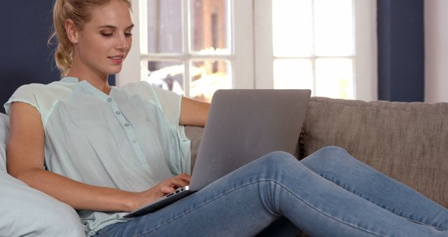 Young woman is working on laptop while sitting on sofa at home. This image is ideal for articles and marketing materials related to remote work, freelance lifestyle, home comfort, or online technology usage. It conveys a sense of productivity and modern living.