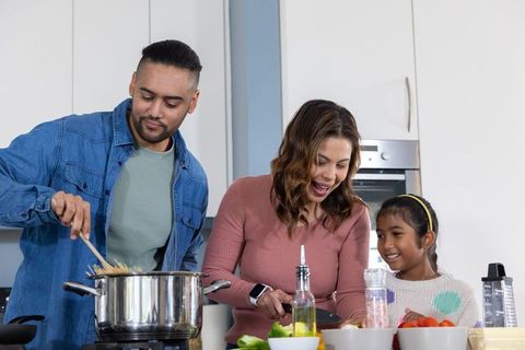 Family cooking together in modern kitchen, father stirring pasta, mother flipping pan, child smiling