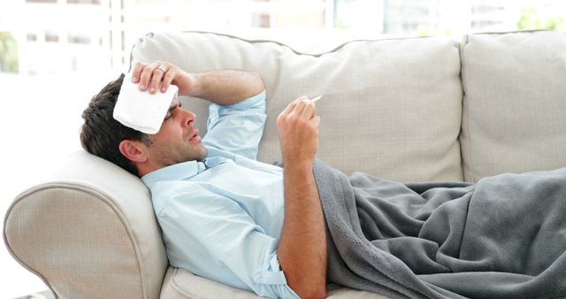 This image shows a man laying on a beige couch, holding a thermometer and using a cold compress on his forehead. He appears to be ill, suggesting he has a fever or the flu. This visual is suitable for use in health blogs, articles about flu season, healthcare promotions, wellness content, and advertising products related to recovery and home care.