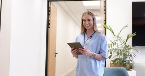 Smiling nurse holding tablet with stethoscope in clinic hallway checking patient records