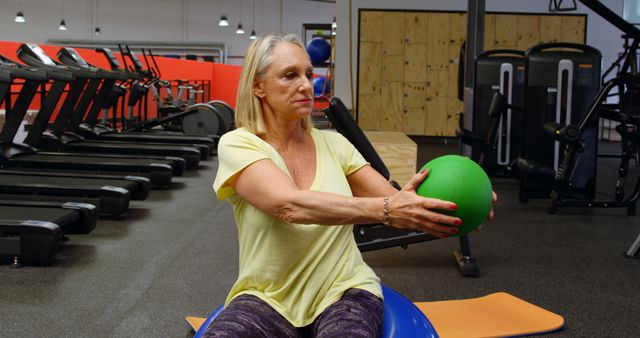 A senior woman is exercising with a green medicine ball in a gym equipped with treadmills and fitness machines. She is sitting on a stability ball while extending the medicine ball in front of her. This image can be used for articles and advertisements about senior fitness, healthy living, exercise routines, and strength training for older adults.