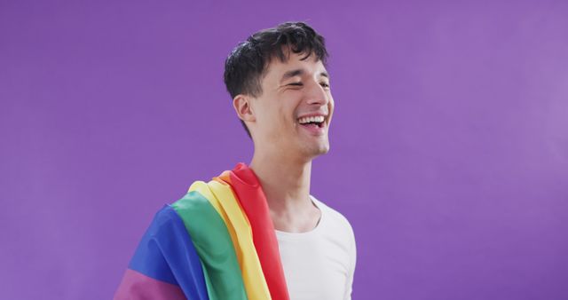 Joyful young man celebrating LGBTQ+ rights with rainbow flag draped over his shoulder against a purple background. Great for content promoting LGBTQ pride, diversity, and inclusivity. Ideal for social media, educational materials, and campaigns advocating human rights and acceptance.