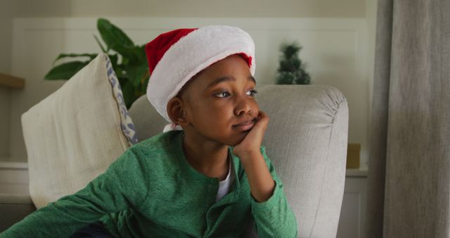 Sad african american boy wearing santa hat sitting on sofa at christmas time. christmas, festivity and tradition at home.