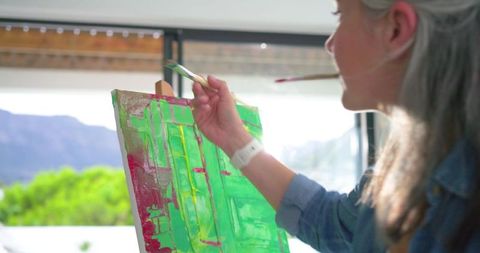Artist painting vibrant magenta and green abstract canvas at sunlit window easel