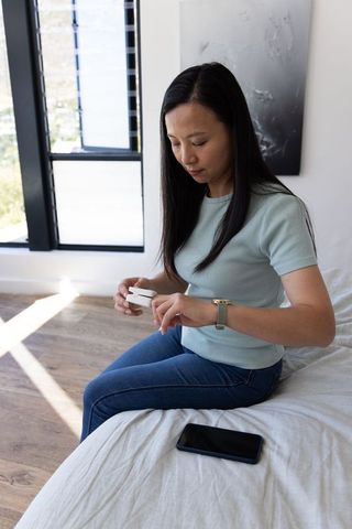 Asian woman sitting on bed inspecting wireless earbud case with smartphone nearby in modern bedroom