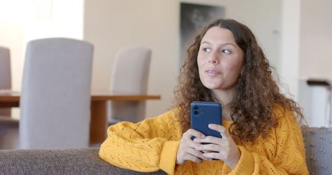 Young woman relaxing on sofa holding smartphone in cozy mustard sweater, looking away