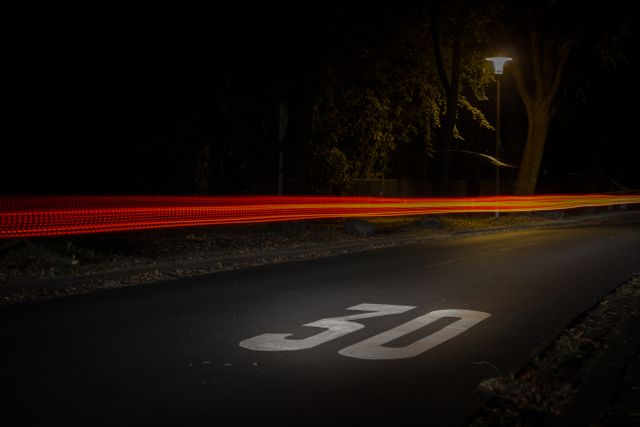 Night scene of a road with a clearly visible speed limit sign of 30 on the pavement. Red light trails from passing cars create dynamic movement, contrasting against the dark background. Trees and a lamp post on the right side illuminate the scene, adding to the ambience. This visual can be used in contexts involving traffic, speed, transportation, or nightlife.