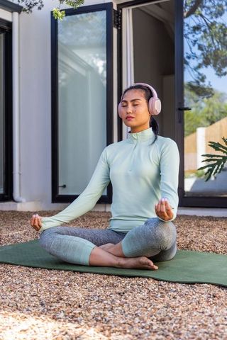 Asian woman meditating outdoors on yoga mat wearing pink headphones practicing mindfulness