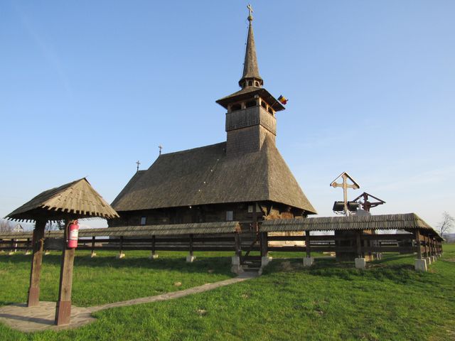 This image depicts an ancient wooden church with a prominent bell tower under a clear blue sky. The structure is surrounded by a wooden fence, emphasizing its historical and cultural heritage. The grassy area around it is green, adding a vibrant touch. This type of image is suitable for content related to cultural heritage, tourism, history, architecture, and religion. It can be used in travel brochures, historical documentaries, educational materials, and religious articles.
