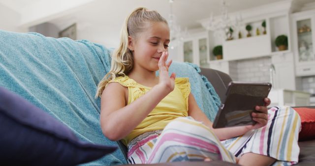 Young girl sitting on a couch, using a tablet for a video call in a bright, modern living room. Ideal for illustrating themes of modern communication, childhood technology use, and staying connected with family and friends. Can be used for advertisements, educational articles, and blogs about kids and technology.
