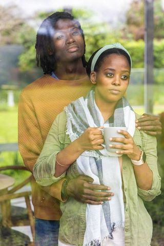 African american couple embracing by window contemplative woman holding warm mug wearing scarf