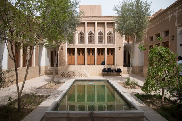 This image features a tranquil courtyard with a calm pool and stunning traditional Persian architecture. Mature trees and well-maintained structures add to its serenity and charm. Ideal for use in travel blogs, heritage articles, and cultural education materials.