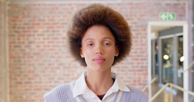 Young woman with afro hairstyle standing confidently in urban building with brick wall in background. The image is well-suited for advertisements emphasizing contemporary lifestyle, fashion editorials, or campaigns focused on diversity and natural beauty.