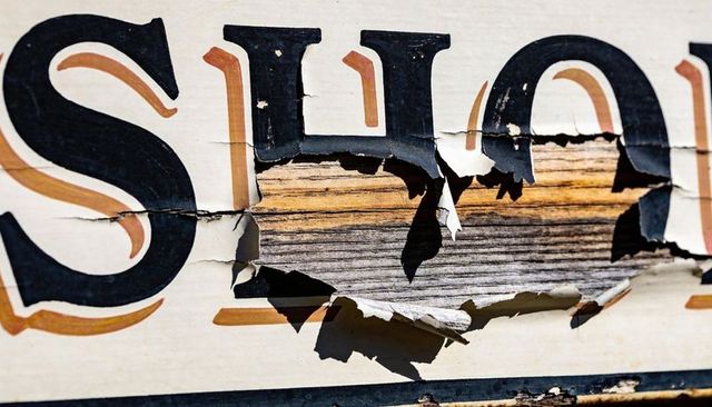 Weathered wooden sign peeling paint revealing woodgrain beneath black serif lettering