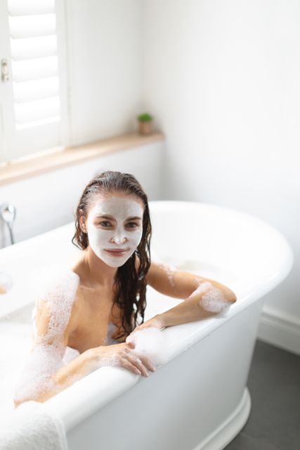 Woman enjoying a relaxing moment in a white bathtub with a face mask applied. Ideal for use in wellness blogs, skincare product advertisements, spa marketing materials, and self-care articles.