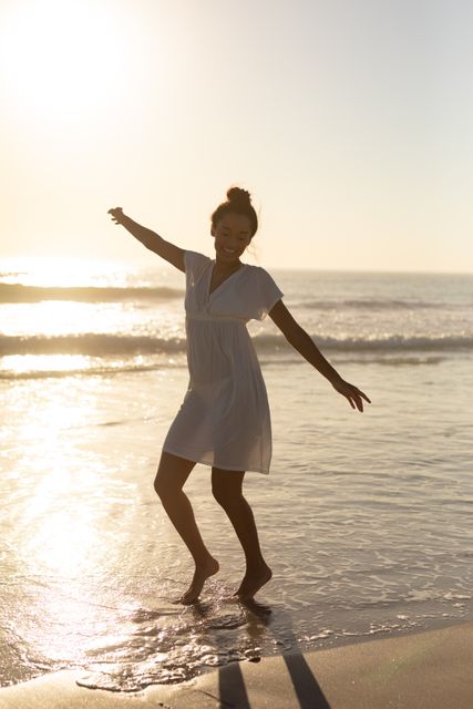 Young woman in white dress dancing joyfully on beach during sunset. Ideal for themes of happiness, freedom, summer vacations, and outdoor lifestyle. Perfect for travel brochures, wellness blogs, and advertisements promoting beach destinations.