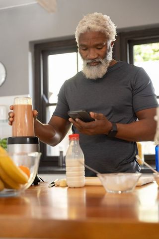 Senior african-american man making smoothie while checking smartphone in modern kitchen