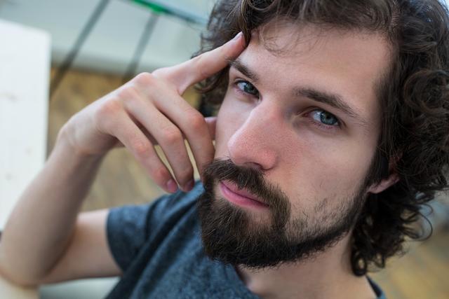 Young male graphic designer with beard and curly hair thinking in office. Ideal for use in articles about creativity, design thinking, modern workplaces, and professional profiles.