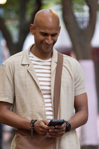Smiling african american man walking using smartphone with crossbody bag in city park