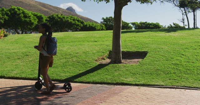 A woman is riding an electric scooter in a park with green trees and a well-manicured lawn. She is wearing a backpack, indicating she may be commuting or enjoying a leisurely ride. This image can be used for promoting eco-friendly transportation, outdoor activities, or urban mobility solutions.