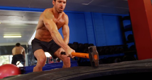 Shirtless man training intensely with a sledgehammer hitting a tire in a gym. Ideal for fitness and strength training advertisements, fitness blogs, gym membership promotions, and motivational workout posters.