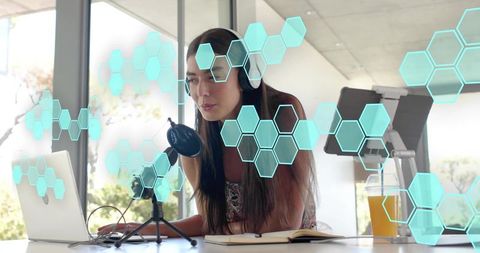 Woman recording podcast at home wearing headphones leaning toward condenser mic with laptop