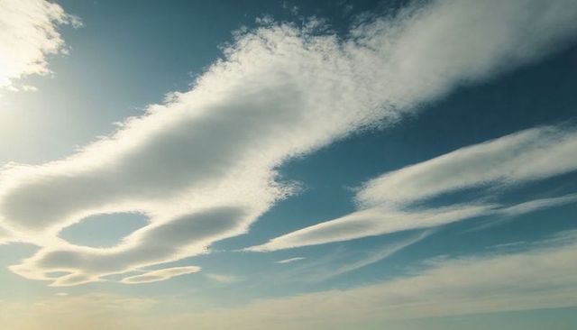 Lenticular clouds revealing rounded holes over vast blue sky with soft sun glare
