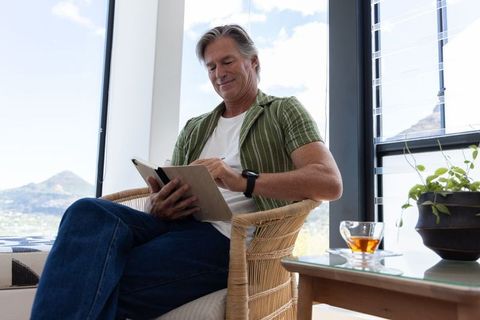 Mature man reading tablet in sunlit wicker chair with mountain view and sipping tea