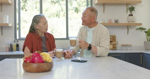 Asian senior couple sharing morning coffee at bright modern kitchen island holding hands